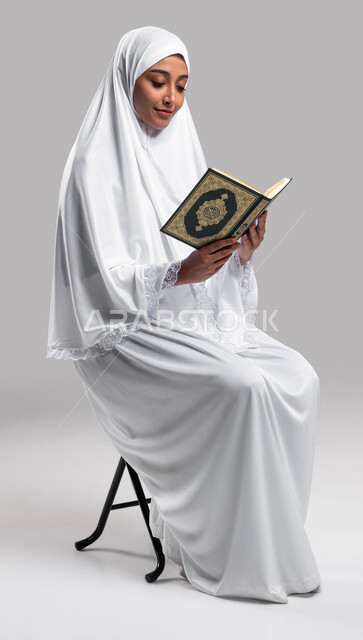 التقرب الى الله بقراءة القرآن الكريم وتدبر معانيه, الاجتهاد في الطاعات في الأشهر المباركة, أداء مناسك الحج والعمرة ، بورتريه لشابة عربية سعودية خليجية تجلس على الكرسي و ترتدي لباس الصلاة و تحمل بيدها كتاب القرآن الكريم ، خلفية رمادية