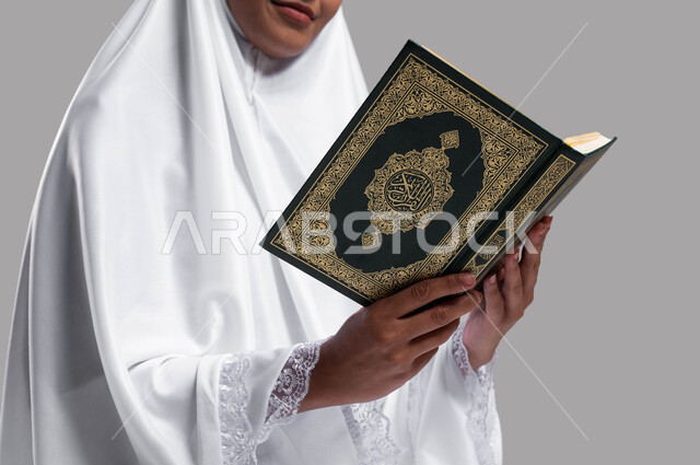 الاجتهاد في العبادة في الأشهر المباركة, أداء مناسك الحج والعمرة ، التقرب الى الله بقراءة القرآن الكريم وتدبر معانيه, بورتريه صورة مقربة لإمرأة عربية سعودية خليجية ترتدي لباس الصلاة و تحمل بيدها كتاب القرآن الكريم ، خلفية رمادية