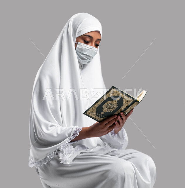 تدبر وقراءة كتاب الله، الالتزام بالإحترازات الوقائية لكورونا, اداء مناسك الحج والعمرة في موسم2023 ، التقرب الى الله بقراءة القرآن, بورتريه لشابة عربية سعودية خليجية ترتدي لباس الصلاة والكمامة الواقية و تحمل بيدها كتاب القرآن الكريم ، خلفية رمادية