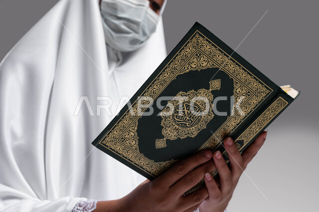 Commitment to the preventive precautions of Corona, performing the rituals of Hajj and Umrah in the 2023 season, drawing closer to God by reading the Qur’an, a portrait of a Saudi Arab Gulf woman wearing a prayer dress and a protective mask and holding in