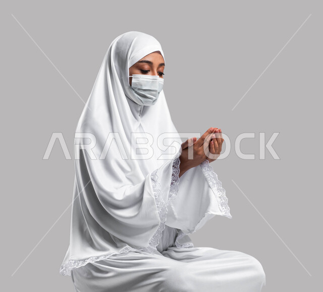 التقرب الى الله بالدعاء والطلب منه, الالتزام بالإجراءات الوقائية لكورونا, بورتريه لإمرأة مسلمة خليجية عربية سعودية ترتدي ثوب الصلاة والكمامة الواقية وترفع يديها للدعاء, ايماءات وتعابير وجه تدل على التضرع الى الله, أداء مناسك الحج والعمرة, خلفية رمادية