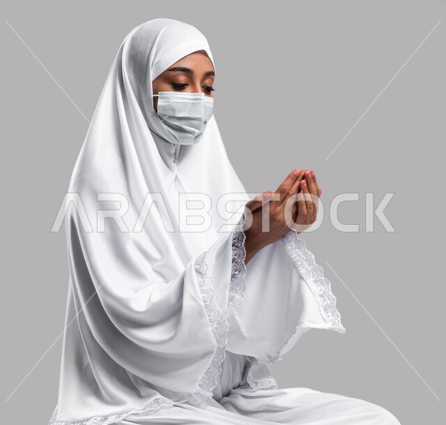 ايماءات وتعابير وجه تدل على التضرع و التمني من الله, الالتزام بالإجراءات الوقائية لفايروس كورونا, بورتريه لإمرأة مسلمة خليجية عربية سعودية ترتدي ثوب الصلاة والكمامة الواقية وترفع يديها للدعاء, أداء مناسك الحج والعمرة, التقرب الى الله بالدعاء, خلفية رمادية