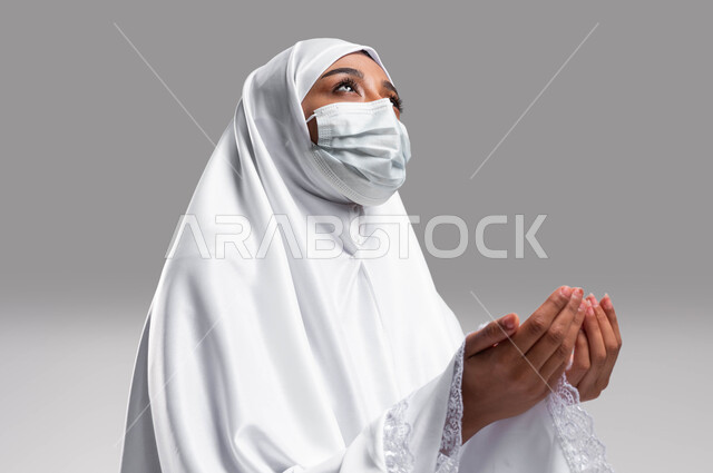 الالتزام بالإجراءات الوقائية لكورونا, بورتريه لإمرأة مسلمة خليجية عربية سعودية ترتدي ثوب الصلاة والكمامة الواقية وترفع يديها للدعاء, ايماءات وتعابير وجه تدل على التضرع الى الله, أداء مناسك الحج والعمرة, التقرب الى الله بالدعاء والطلب منه, خلفية رمادية