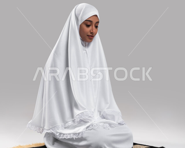 أداء مناسك الحج والعمرة في موسم الحج 2023، الجلوس على سجادة الصلاة والتقرب الى الله بأداء الطاعات، بورتريه لامرأة مسلمة عربية سعودية خليجية ترتدي ثياب الصلاة وتؤدي فريضة الصلاة، خلفية رمادية