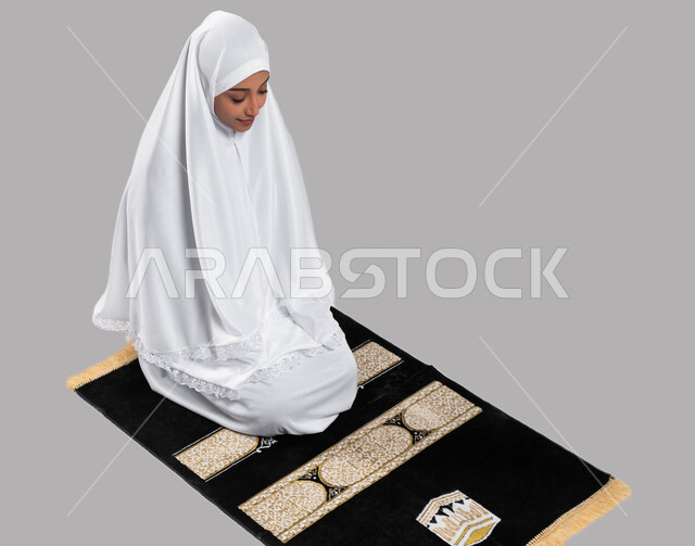 الجلوس على سجادة الصلاة والتقرب الى الله بأداء الطاعات، بورتريه لشابة مسلمة عربية سعودية خليجية ترتدي ثياب الصلاة وتؤدي فريضة الصلاة، أداء مناسك الحج والعمرة في موسم الحج 2023، خلفية رمادية