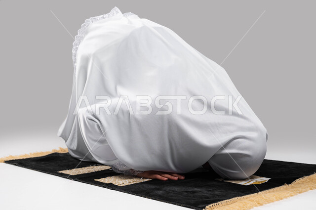 الصلاة و التضرع الى الله, السجود على سجادة الصلاة, بورتريه لإمرأة عربية سعودية خليجية تلبس زي الاحرام و تؤدي فريضة الصلاة ، أداء مناسك الحج والعمرة ، التقرب الى الله بأداء العبادات والطاعات, خلفية رمادية