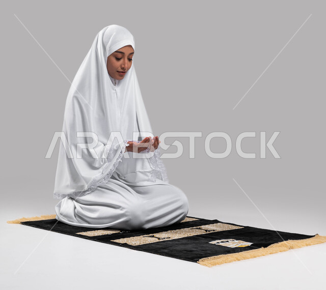 آداء مناسك الحج والعمرة، إيماءات وتعابير وجه تدل على التضرع الى الله, الجلوس على سجادة الصلاة والدعاء والعباده والتقرب الى الله، بورتريه لشابة مسلمة عربية سعودية خليجية ترتدي ملابس الاحرام وترفع يديها للدعاء، موسم الحج 2023، خلفية رمادية