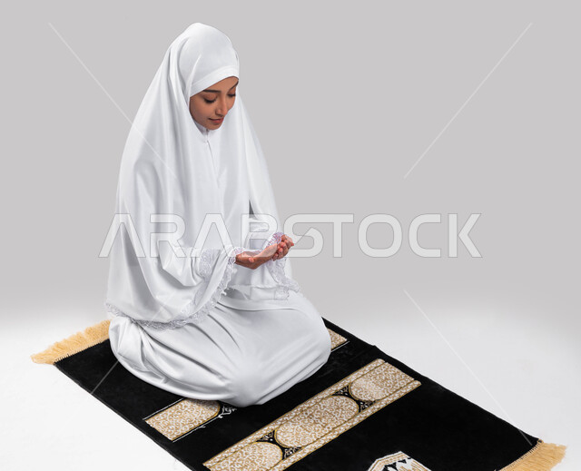 موسم الحج 2023، الجلوس على سجادة الصلاة والدعاء والعباده والتقرب الى الله، آداء مناسك الحج والعمرة، بورتريه لشابة مسلمة عربية سعودية خليجية ترتدي ملابس الاحرام وترفع يديها للدعاء، خلفية رمادية