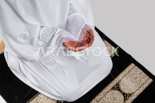 الجلوس على سجادة الصلاة والدعاء والعباده والتقرب الى الله، آداء مناسك الحج والعمرة، بورتريه صورة مقربة ليدين شابة مسلمة عربية سعودية خليجية ترتدي ملابس الاحرام وترفع يديها للدعاء، موسم الحج 2023، خلفية رمادية