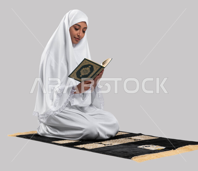 إيماءات وتعابير وجه تعبر عن تدبر آيات كتاب الله, التقرب الى الله بقراءة القرآن, بورتريه لإمرأة عربية سعودية خليجية ترتدي لباس الاحرام تجلس على سجادة الصلاة و تحمل بيدها كتاب القرآن الكريم ، اداء مناسك الحج والعمرة ، خلفية رمادية