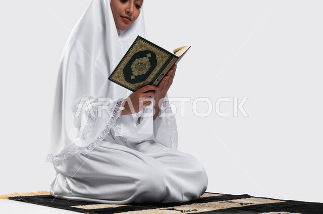 إيماءات وجه تدل على تدبر كتاب الله, التقرب الى الله بقراءة القرآن, بورتريه لإمرأة عربية سعودية خليجية ترتدي لباس الاحرام تجلس على سجادة الصلاة و تحمل بيدها كتاب القرآن الكريم ، موسم الحج 2023، خلفية رمادية