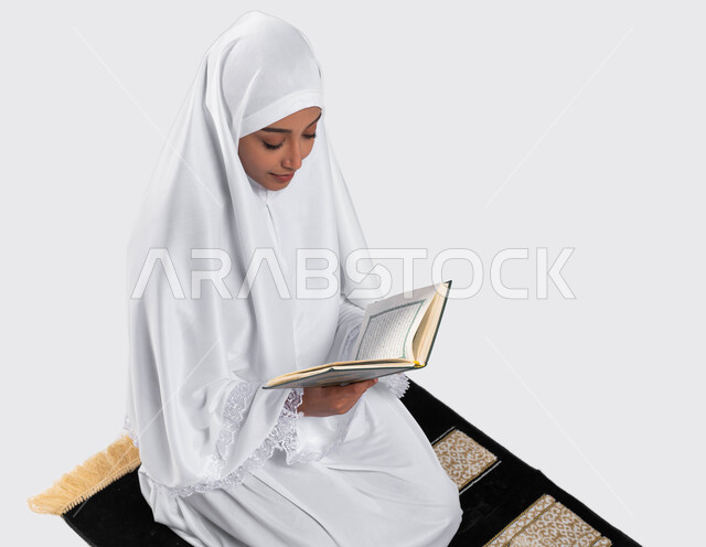 اداء مناسك الحج والعمرة ، التقرب الى الله بقراءة القرآن, بورتريه لإمرأة عربية سعودية خليجية ترتدي لباس الاحرام تجلس على سجادة الصلاة و تحمل بيدها كتاب القرآن الكريم ، تدبر وقراءة كتاب الله ، موسم الحج 2023، خلفية رمادية