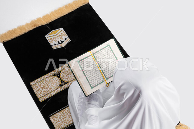 التقرب الى الله بقراءة القرآن, بورتريه بزاوية علوية عامودية لإمرأة عربية سعودية خليجية ترتدي لباس الاحرام تجلس على سجادة الصلاة و تحمل بيدها كتاب القرآن الكريم ، تدبر وقراءة كتاب الله ، موسم الحج 2023، اداء مناسك الحج والعمرة ، خلفية رمادية