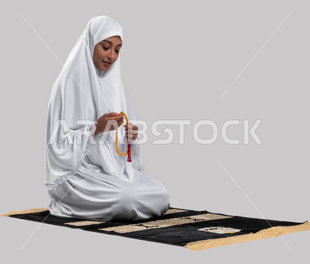 موسم الحج 2023، قراءة أذكار ما بعد الصلاة,آداء مناسك الحج والعمرة، بورتريه لإمرأة مسلمة عربية سعودية خليجية ترتدي ملابس الاحرام و تحمل بيدها سبحة، الجلوس على سجادة الصلاة والقيام بالتسبيح وقراءة الأذكار، خلفية رمادية