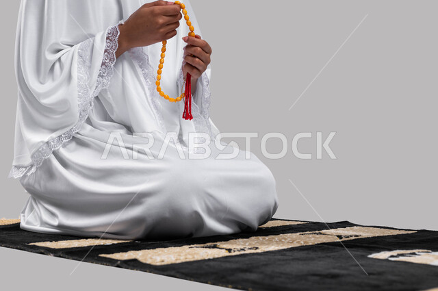 الجلوس على سجادة الصلاة والقيام بالتسبيح وقراءة الأذكار، قراءة أذكار ما بعد الصلاة,آداء مناسك الحج والعمرة، بورتريه لشابة مسلمة عربية سعودية خليجية ترتدي ملابس الاحرام و تحمل بيدها سبحة، موسم الحج 2023، خلفية رمادية