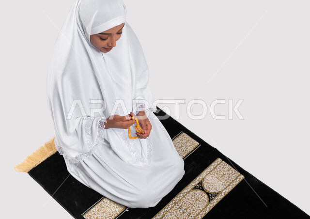 آداء مناسك الحج والعمرة، قراءة أذكار ما بعد الصلاة, بورتريه لإمرأة مسلمة عربية سعودية خليجية ترتدي ملابس الاحرام و تحمل بيدها سبحة، الجلوس على سجادة الصلاة والقيام بالتسبيح وقراءة الأذكار، موسم الحج 2023، خلفية رمادية