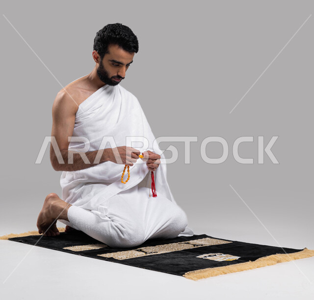 آداء مناسك الحج والعمرة, قراءة أذكار ما بعد الصلاة, بورتريه لرجل مسلم عربي سعودي خليجي يرتدي ملابس الاحرام ويحمل بيده سبحة، الجلوس على سجادة الصلاة والقيام بالتسبيح وقراءة الأذكار، موسم الحج 2023، خلفية رمادية