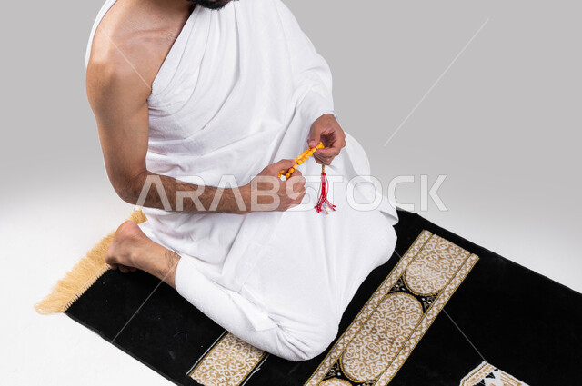 قراءة أذكار ما بعد الصلاة,آداء مناسك الحج والعمرة، بورتريه لرجل مسلم عربي سعودي خليجي يرتدي ملابس الاحرام ويحمل بيده سبحة، الجلوس على سجادة الصلاة والقيام بالتسبيح وقراءة الأذكار، موسم الحج 2023، خلفية رمادية
