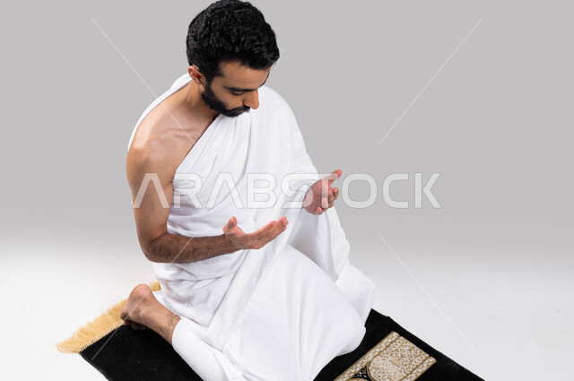 آداء مناسك الحج والعمرة، بورتريه لشاب مسلم عربي سعودي خليجي يرتدي ملابس الاحرام ويرفع يديه للدعاء، الجلوس على سجادة الصلاة والدعاء والعباده والتقرب الى الله، موسم الحج 2023، خلفية رمادية