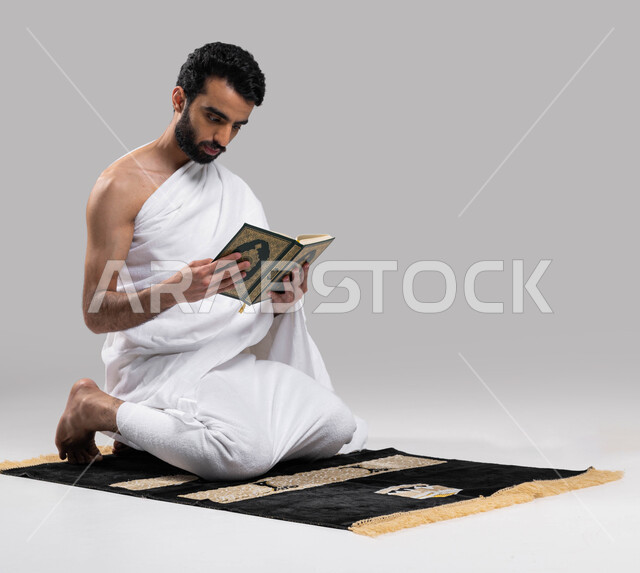 ايماءات تعبر عن التمعن بآيات كتاب الله, التقرب الى الله بقراءة القرآن, بورتريه لرجل عربي سعودي خليجي يرتدي لباس الاحرام يجلس على سجادة الصلاة و يحمل بيده كتاب القرآن الكريم ، موسم الحج 2023، اداء مناسك الحج والعمرة ، خلفية رمادية