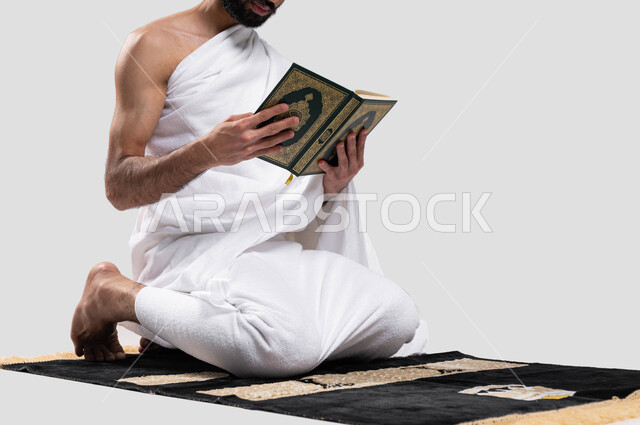 مفهوم التمعن بآيات وسور كتاب الله, التقرب الى الله بقراءة القرآن, بورتريه لرجل عربي سعودي خليجي يرتدي لباس الاحرام يجلس على سجادة الصلاة و يحمل بيده كتاب القرآن الكريم ، موسم الحج 2023، اداء مناسك الحج والعمرة ، خلفية رمادية