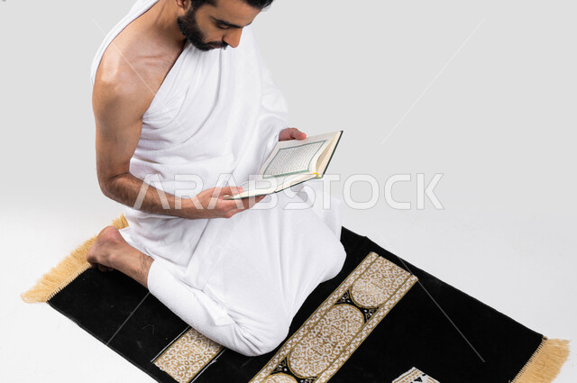 قراءة كتاب الله ، التقرب الى الله بقراءة القرآن, بورتريه لرجل عربي سعودي خليجي يرتدي لباس الاحرام يجلس على سجادة الصلاة و يحمل بيده كتاب القرآن الكريم ، قراءة كتاب الله ، موسم الحج 2023، اداء مناسك الحج والعمرة ، خلفية رمادية