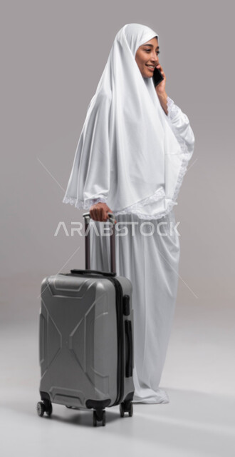 إيماءات تدل على السعادة باستخدام الهاتف الجوال والاستعداد للسفر لآداء مناسك الحج والعمرة، بورتريه صورة جانبية لشابة مسلمة خليجية عربية سعودية تلبس ملابس الاحرام تمسك بيدها حقيبة السفر و تجري مكالمة هاتفية, موسم الحج 2023, صورة الجسم بالطول الكامل, خلفية ر