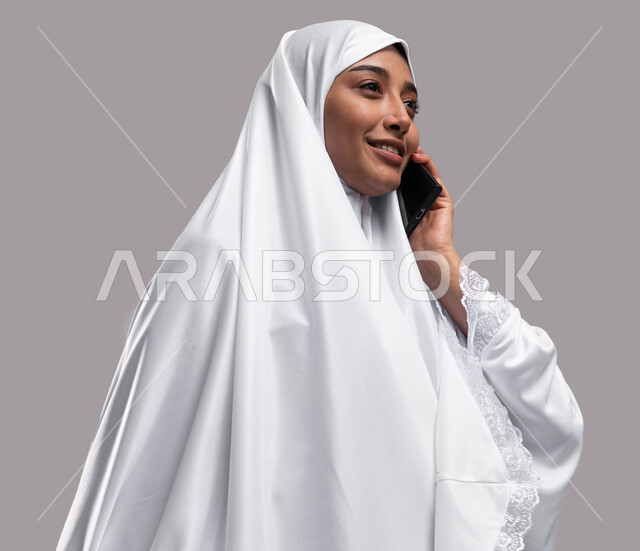 آداء مناسك الحج والعمرة، استخدام الهاتف المحمول، بورتريه صورة جانبية لإمراة مسلمة خليجية عربية سعودية ترتدي ملابس الاحرام و تجري مكالمة هاتفية، التواصل الاجتماعي والعلاقات الاجتماعية، موسم الحج 2023, خلفية رمادية