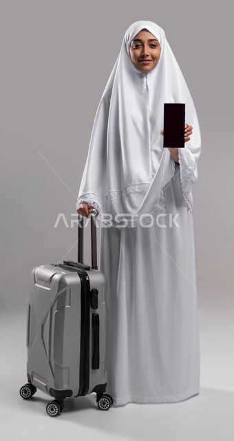 الاستعداد للسفر لأداء فريضة الحج, التقرب الى الله بأداء الطاعات, بورتريه لإمراة مسلمة خليجية عربية سعودية ترتدي ملابس الاحرام وتمسك بيدها حقيبة السفر و الهاتف المحمول بشاشة سوداء فارغة، آداء مناسك الحج والعمرة2023، صورة الجسم بالطول الكامل, خلفية رمادية