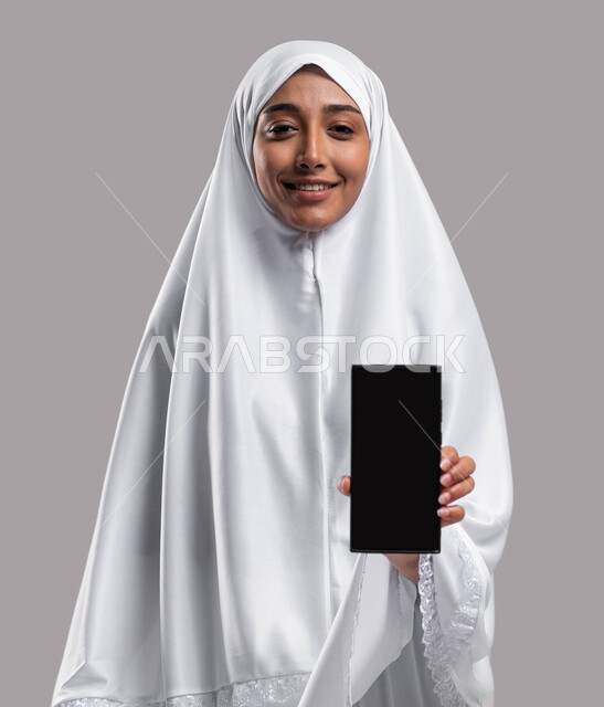 ايماءات وجه تدل على السعادة, التقرب الى الله بأداء الطاعات, بورتريه إمراة مسلمة خليجية عربية سعودية ترتدي ملابس الاحرام تحمل بيدها الهاتف المحمول بشاشة سوداء فارغة، موسم الحج 2023، آداء مناسك الحج والعمرة، خلفية رمادية
