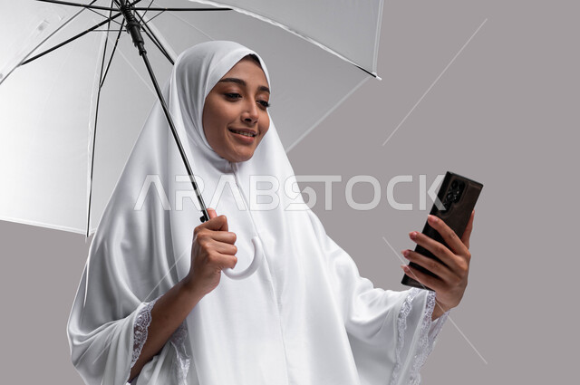 ايماءات تدل على السعادة بإجراء مكالمة فيديو والتواصل مع العائلة والأصدقاء، موسم الحج 2023, بورتريه لإمراة مسلمة خليجية عربية سعودية ترتدي ملابس الاحرام تحمل بيدها مظلة وتجري مكالمة هاتفية، آداء مناسك الحج والعمرة، خلفية رمادية