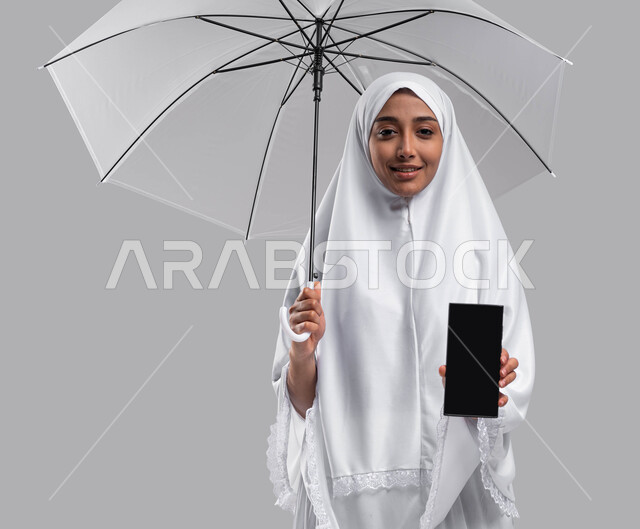 التقرب الى الله بأداء الطاعات, بورتريه إمراة مسلمة خليجية عربية سعودية ترتدي ملابس الاحرام وتحمل بيدها مظلة  و الهاتف المحمول بشاشة سوداء فارغة، موسم الحج 2023، آداء مناسك الحج والعمرة، الركن الخامس من أركان الاسلام, خلفية رمادية