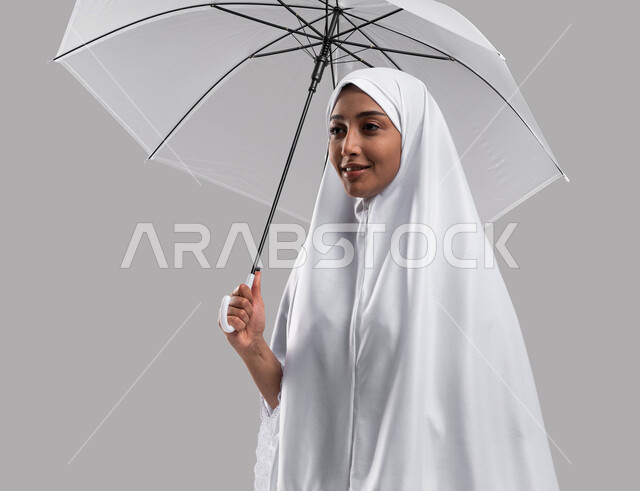 إيماءات تدل على السعادة بأداء مناسك الحج و العمرة, بورتريه إمراة مسلمة خليجية عربية سعودية ترتدي ملابس الاحرام وتحمل بيدها مظلة , موسم الحج 2023، الركن الخامس من أركان الاسلام، خلفية رمادية