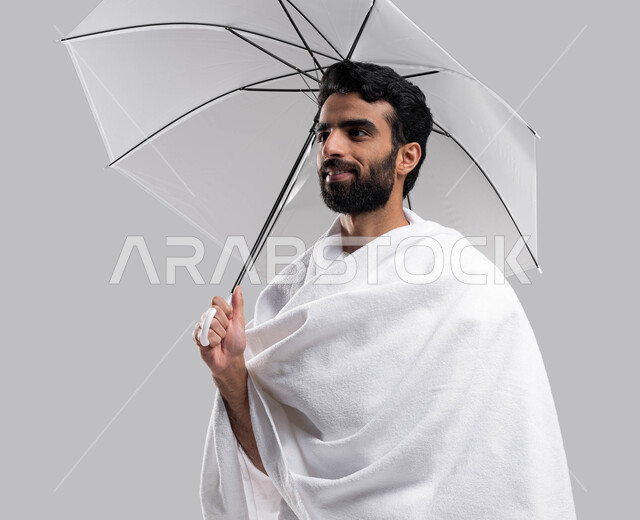 The Fifth Pillar of Islam, a portrait of a Saudi Arabian Gulf Muslim man wearing ihram clothes and holding an umbrella in his hand, Hajj season 2023, gestures indicating happiness in performing Hajj and Umrah rituals, gray background