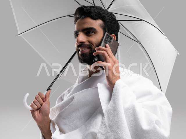 ايماءات تدل على السعادة بالتواصل مع العائلة والأصدقاء، بورتريه لشاب مسلم خليجي عربي سعودي يلبس ملابس الاحرام يجري مكالمة هاتفية ويحمل بيده مظلة رمادية، التواصل الاجتماعي والعلاقات الاجتماعية، آداء مناسك الحج والعمرة، موسم الحج 2023، خلفية رمادية