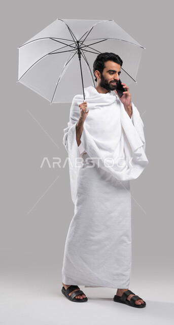  التواصل الاجتماعي والعلاقات الاجتماعية، ايماءات تدل على السعادة بالتواصل مع العائلة والأصدقاء، بورتريه لرجل  مسلم خليجي عربي سعودي يرتدي ملابس الاحرام يجري مكالمة هاتفية ويحمل بيده مظلة رمادية، آداء مناسك الحج والعمرة، صورة الجسم بالطول الكامل, خلفية رما