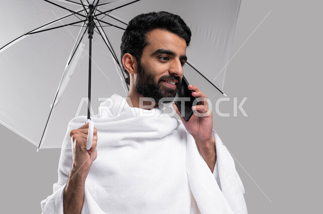 ايماءات تدل على السعادة بالتواصل مع العائلة والأصدقاء، بورتريه لرجل  مسلم خليجي عربي سعودي يرتدي ملابس الاحرام يجري مكالمة هاتفية ويحمل بيده مظلة رمادية، التواصل الاجتماعي والعلاقات الاجتماعية، آداء مناسك الحج والعمرة، موسم الحج 2023، خلفية رمادية