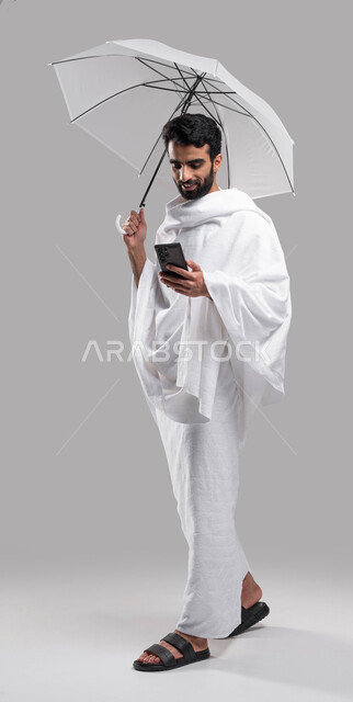 التقرب الى الله بآداء مناسك الحج والعمرة، الركن الخامس من أركان الإسلام, بورتريه لشاب مسلم خليجي عربي سعودي يرتدي ملابس الاحرام ويحمل بيده شمسية رمادية ويقوم بتصفح الهاتف الجوال، صورة الجسم بالطول الكامل, خلفية رمادية