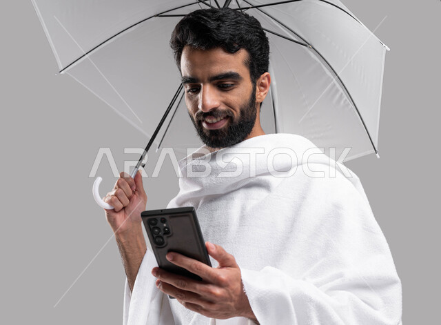 التقرب الى الله بآداء مناسك الحج والعمرة، بورتريه لشاب مسلم خليجي عربي سعودي يرتدي ملابس الاحرام ويحمل بيده شمسية رمادية ويقوم بتصفح الهاتف الجوال، موسم الحج 2023 ، خلفية رمادية