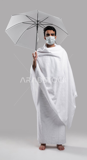 ارتداء الكمامة الواقية, موسم الحج 2023 والوقاية من فايروس كورونا، بورتريه لرجل مسلم خليجي عربي سعودي يرتدي ملابس الاحرام ويحمل بيده مظلة رمادية، صورة الجسم بالطول الكامل, خلفية رمادية