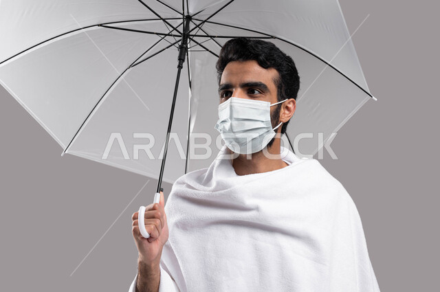 Pilgrims wearing a protective mask, the Hajj season 2023, and prevention from the Corona virus, a close-up portrait of a young Saudi Arabian Gulf Muslim wearing Ihram clothes and holding a gray umbrella in his hand, performing the rituals of Hajj and Umra