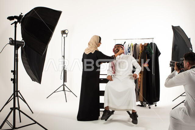 تصوير اعلاني تسويقي، مصور محترف عربي سعودي خليجي يقوم بالتقاط صور فوتوغرافية لامرأة تقوم بتجميل وتحضير العارض، بورتريه لجلسة تصوير في الاستوديو، كاميرات ومعدات متطورة، خلف الكواليس، عبايات نسائية خليجية، خلفية بيضاء