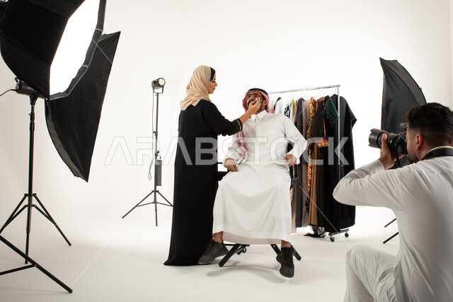 امرأة تقوم بتجميل وتحضير العارض لتصوير اعلاني تسويقي، بورتريه لجلسة تصوير في الاستوديو، مصور محترف عربي سعودي خليجي يقوم بالتقاط صور فوتوغرافية، كاميرات ومعدات متطورة، خلف الكواليس، عبايات نسائية خليجية، خلفية بيضاء