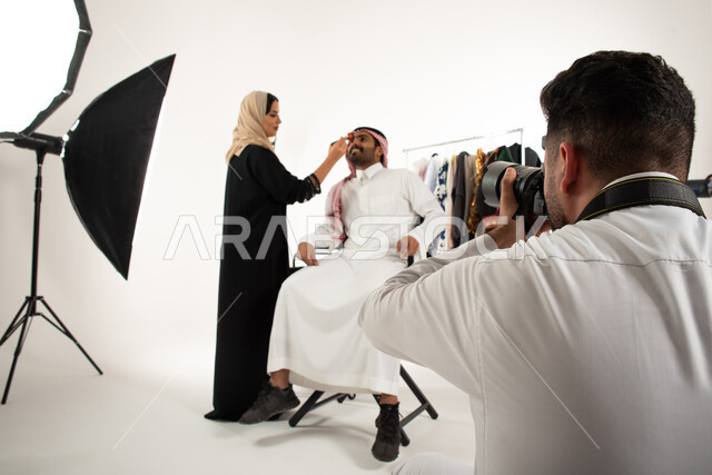خلف الكواليس، عبايات نسائية خليجية، بورتريه لجلسة تصوير في الاستوديو، مصور محترف عربي سعودي خليجي يقوم بالتقاط صور فوتوغرافية لامرأة تقوم بتجميل وتحضير العارض لتصوير اعلاني تسويقي، كاميرات ومعدات متطورة، خلفية بيضاء