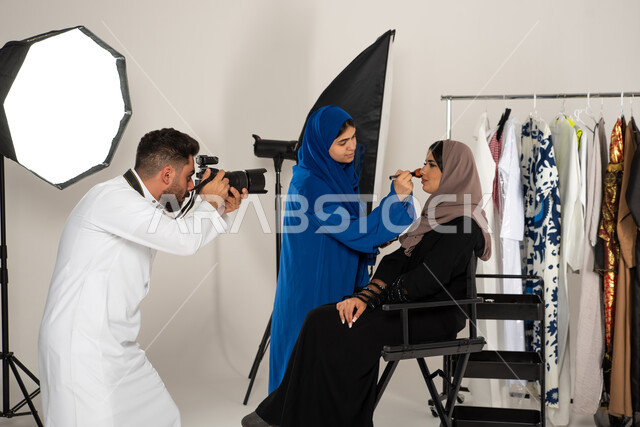 تصوير اعلاني تسويقي، امرأة تقوم بتجميل عارضة ازياء وموديلات عصرية، بورتريه لجلسة تصوير في الاستوديو، مصور عربي سعودي خليجي يقوم بالتقاط صور فوتوغرافية، كاميرات ومعدات متطورة، خلف الكواليس، عبايات نسائية خليجية، خلفية بيضاء