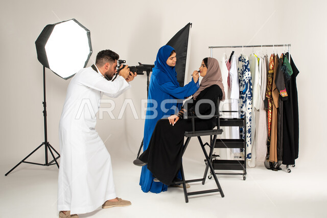 مصور عربي سعودي خليجي يقوم بالتقاط صور فوتوغرافية لامرأة تقوم بتجميل عارضة ازياء وموديلات عصرية، بورتريه لجلسة تصوير في الاستوديو، تصوير اعلاني تسويقي، كاميرات ومعدات متطورة، خلف الكواليس، عبايات نسائية خليجية، خلفية بيضاء
