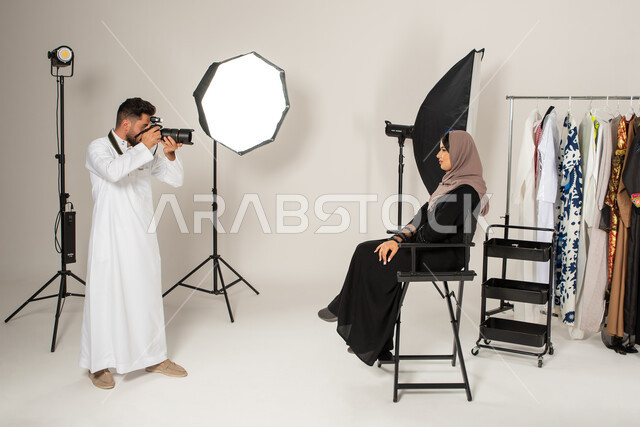عارضة ازياء وموديلات عصرية، خلف الكواليس، بورتريه لجلسة تصوير في الاستوديو، مصور عربي سعودي خليجي يقوم بالتقاط صور فوتوغرافية لامرأة تجلس على الكرسي، عبايات نسائية خليجية، كاميرات ومعدات متطورة، تصوير اعلاني تسويقي، خلفية بيضاء