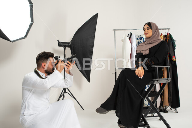 عارضة ازياء وموديلات عصرية، بورتريه لجلسة تصوير في الاستوديو، مصور عربي سعودي خليجي يقوم بالتقاط صور فوتوغرافية لامرأة تجلس على الكرسي، عبايات نسائية خليجية، كاميرات ومعدات متطورة، خلف الكواليس، تصوير اعلاني تسويقي، خلفية بيضاء