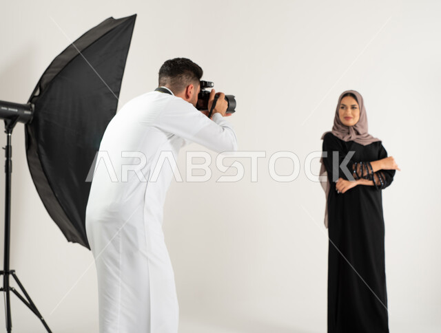 كاميرات ومعدات متطورة، امرأة ترتدي عباءة نسائية خليجية، بورتريه لجلسة تصوير في الاستوديو، مصور محترف عربي سعودي خليجي يقوم بالتقاط صور فوتوغرافية لعارضة أزياء وموديلات عصرية، تصوير اعلاني تسويقي، خلف الكواليس، خلفية بيضاء