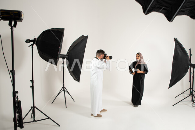 عارضة أزياء وموديلات عصرية، امرأة ترتدي عباءة نسائية خليجية، بورتريه لجلسة تصوير في الاستوديو، مصور محترف عربي سعودي خليجي يقوم بالتقاط صور فوتوغرافية، كاميرات ومعدات متطورة، تصوير اعلاني تسويقي، خلف الكواليس، خلفية بيضاء